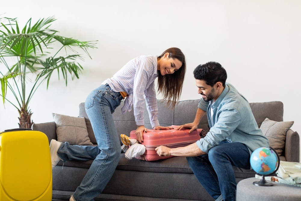 Preparation for vacation. Happy spouses packing and trying to close suitcase on sofa, preparing for travel at home. Vacation, family travel together and positive emotions